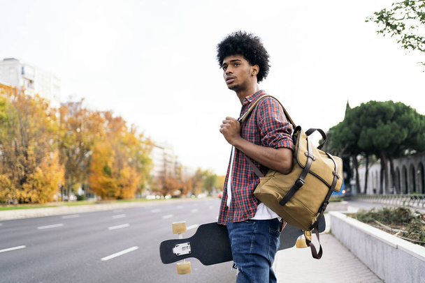 Stock photo of young afro boy walking in the city and carrying his longboard. - Foto, Imagen