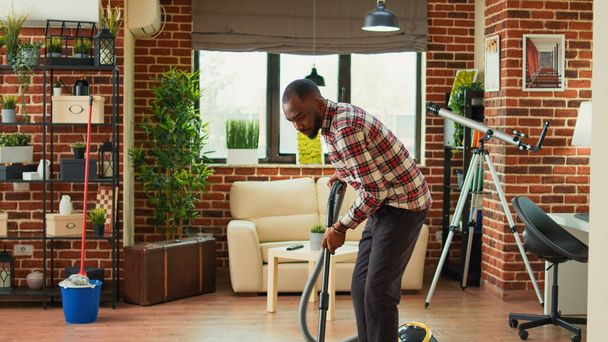 African american partner using vacuum to clean dust in living room, doing spring cleaning work together at home. Young man vacuuming apartment floors and helping with housework. - Foto, Imagen