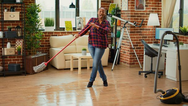 Modern housewife doing dance moves in living room and mopping wooden floors, listening to music and enjoying spring cleaning. Happy person singing and using mop to clean at home. - Foto, imagen