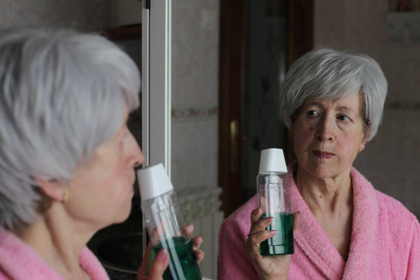 close-up portrait of mature woman with bottle of mouthwash in front of mirror in bathroom - Foto, Imagen