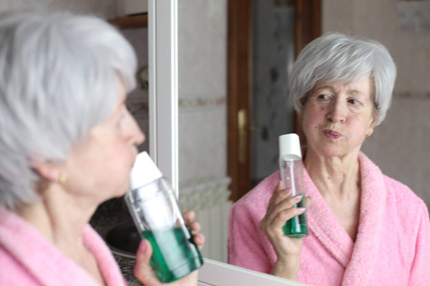 close-up portret van volwassen vrouw met fles mondwater in de voorkant van spiegel in de badkamer - Foto, afbeelding