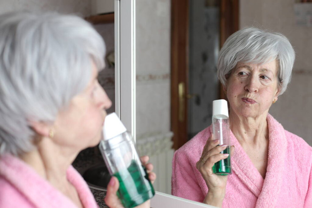 close-up portrait of mature woman with bottle of mouthwash in front of mirror in bathroom - Foto, Bild