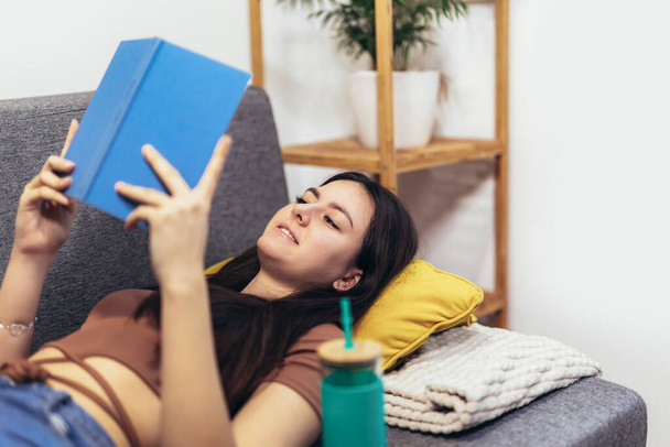 Dospívající dívka ležící na gauči a čte si knihu. Moderní teenager doma. Teen literatura koncept. - Fotografie, Obrázek