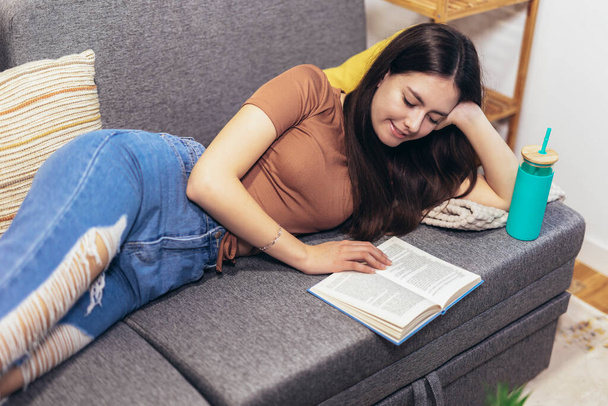 Adolescente allongée sur le canapé et lit un livre. Adolescent moderne à la maison. Concept de littérature pour adolescents. - Photo, image