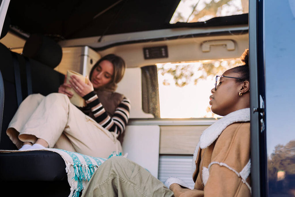 two young friends chilling and reading during a road trip in camper van, concept of travel adventure and female friendship - Foto, Imagen