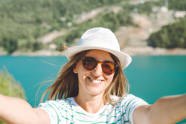 Woman taking selfie photo with wide angle front camera of mountains lake background. Traveler female having fun on the blue lake outdoors travel adventure vacation.  - Photo, Image