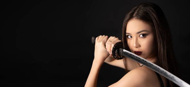 Portret van een mooi Aziatisch meisje met een samoerai katana zwaard op zwarte achtergrond. Banner met kopieerruimte. - Foto, afbeelding