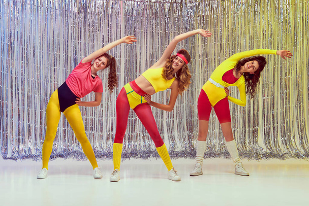 Sportive, active, young women in colorful, comfortable sportswear training, doing stretching and aerobics exercises. Slim fit body. Concept of retro fitness, sport, 80s fashion, beauty, health - 写真・画像