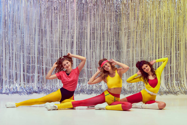 Three stylish young girls in colorful sportswear, bodysuits and leggings training, doing stretching and aerobics exercises. Concept of retro fitness, sport, 80s fashion, beauty, health - Photo, image