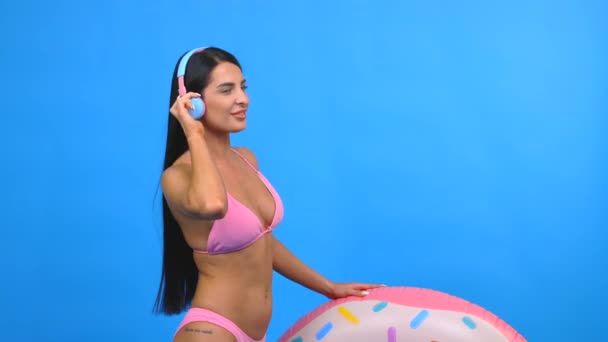 Happy young woman wearing in pink swimsuit listening music in headphones and holding donut inflatable ring, dancing and singing song and having fun isolated on blue wall background - Footage, Video