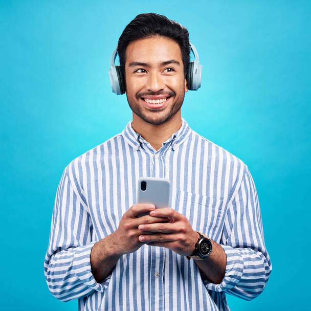 Słuchawki, muzyka i myślący człowiek z telefonem w studio do streamingu, audio i subskrypcji na niebieskim tle. Radio, uśmiech i szczęśliwy azjatycki mężczyzna online podcast, utwór i aplikacja internetowa. - Zdjęcie, obraz