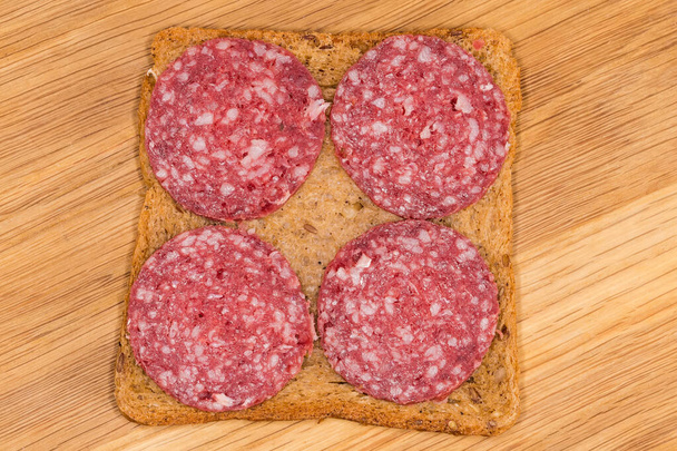 Ανοιχτό σάντουιτς φτιαγμένο με πολύσπορο ψωμί και φέτες από το αποξηραμένο λουκάνικο σε ξύλινη επιφάνεια κοπής, top view close-up - Φωτογραφία, εικόνα