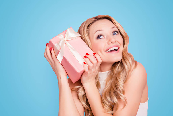 Portrait of joyful cheerful girlfriend having packed gift box in hands trying to know what inside, holding case near ear isolated on pink background. - Fotó, kép