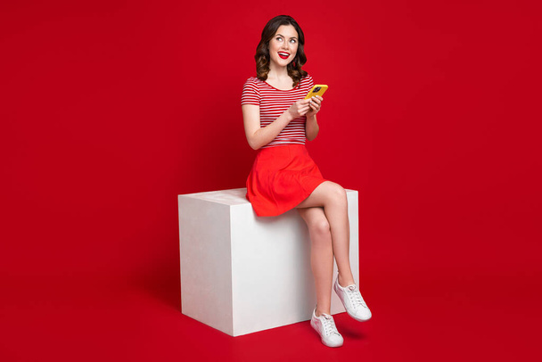 Full body photo of slim lovely lady sit podium use smart phone look empty space isolated on red color background. - Foto, Bild