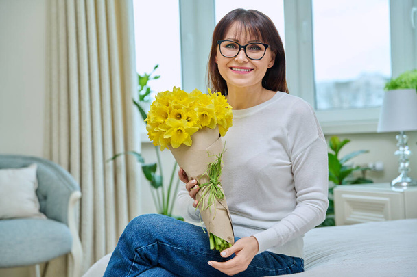 Middle aged attractive smiling woman with bouquet of yellow flowers looking at camera sitting on couch at home, room interior background. Holiday, beauty, emotion, nature, 40s people concept - 写真・画像