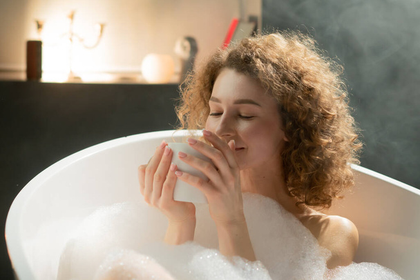Young relaxed woman lying in the bath enjoys the smell of fragrant tea. Attractive woman lies in a bath with foam and holds a cup of hot tea in her hands. High quality photo - Foto, Imagen