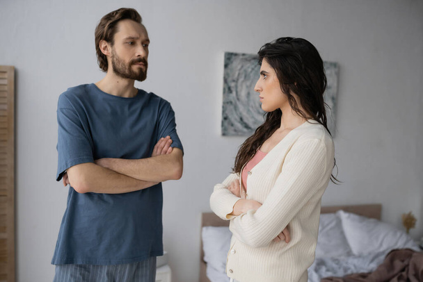 Offended woman crossing arms and looking at bearded boyfriend in pajama in bedroom  - Fotografie, Obrázek