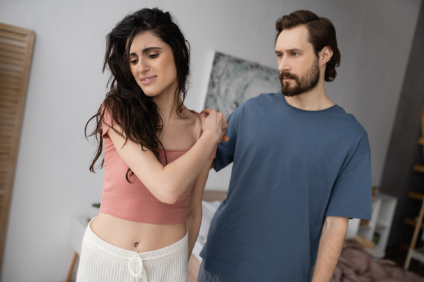 Bearded man touching shoulder of stressed girlfriend in pajama in bedroom - Photo, Image
