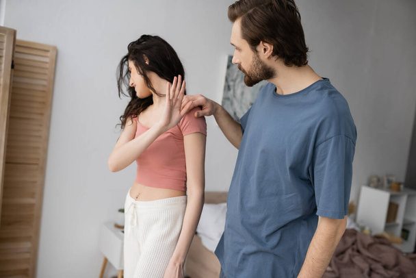 Bearded man calming offended girlfriend showing stop gesture in bedroom  - Photo, Image