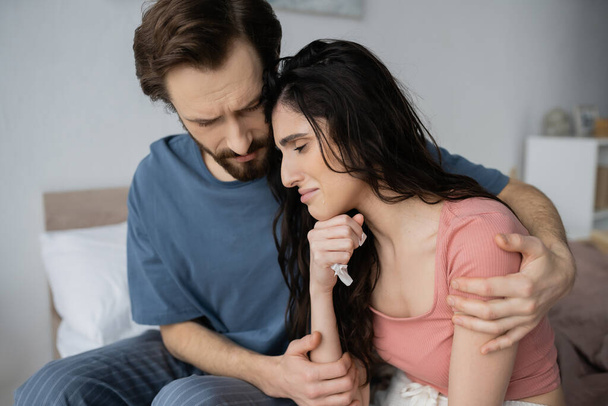 Hombre empático en pijama calmando a su novia llorando con servilleta en el dormitorio  - Foto, Imagen