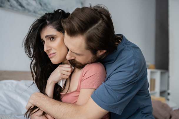 Bärtiger Mann umarmt traurige Freundin mit Serviette im verschwommenen Schlafzimmer  - Foto, Bild