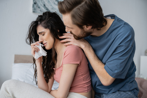 Caring man in pajama hugging crying girlfriend with napkin in bedroom  - Fotografie, Obrázek