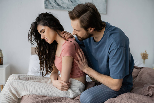 Bearded man calming disappointed girlfriend in pajama in bedroom  - Фото, изображение