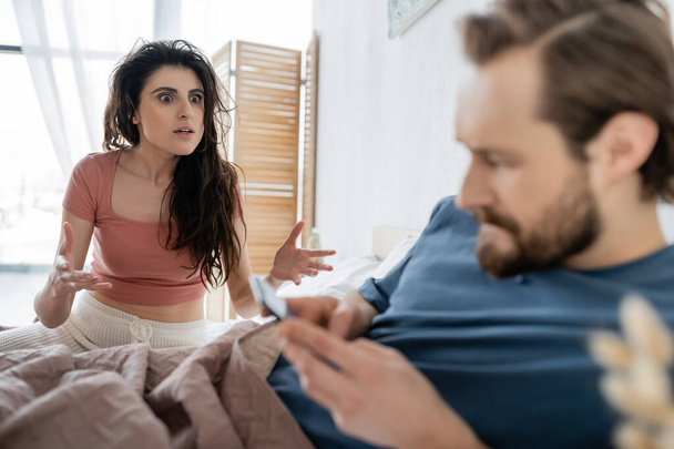 Annoyed woman in pajama quarrelling at blurred boyfriend with smartphone on bed  - Foto, imagen