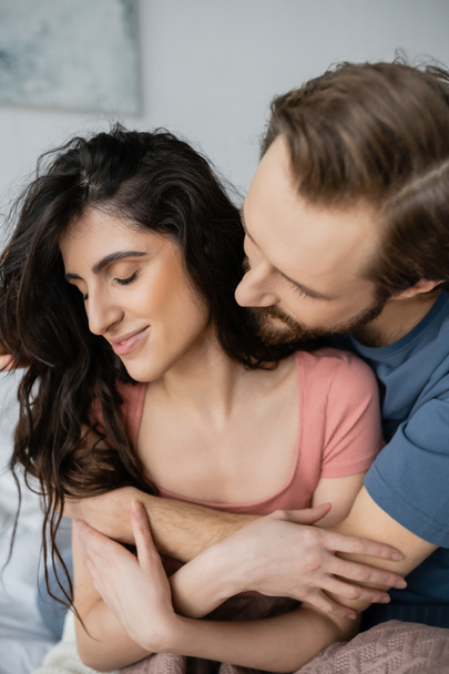 Homem barbudo abraçando sorridente morena namorada no quarto  - Foto, Imagem