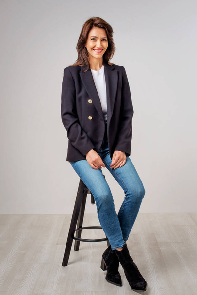 Studio portrait of attractive woman wearing blazer and blue jeans while sitting at isolated white background. Full length shot. Copy space.  - 写真・画像