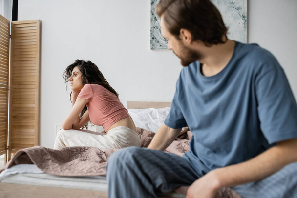 Indifferent woman in pajama sitting on bed near blurred boyfriend at home  - Фото, изображение