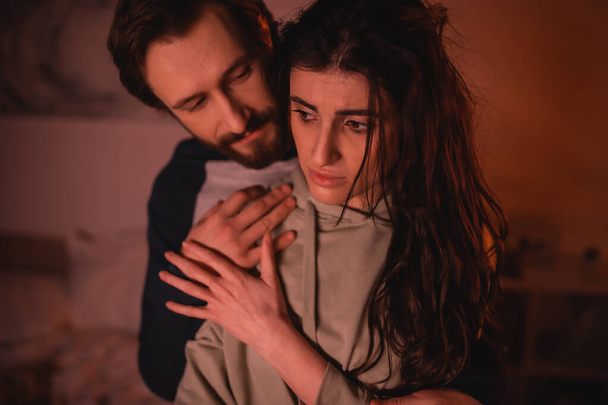 Bärtiger Mann umarmt traurige Freundin abends zu Hause  - Foto, Bild
