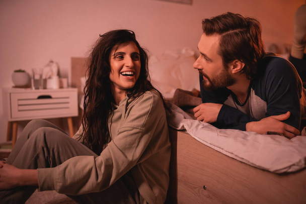 Bearded man talking to cheerful girlfriend in bedroom in evening  - Foto, Imagen