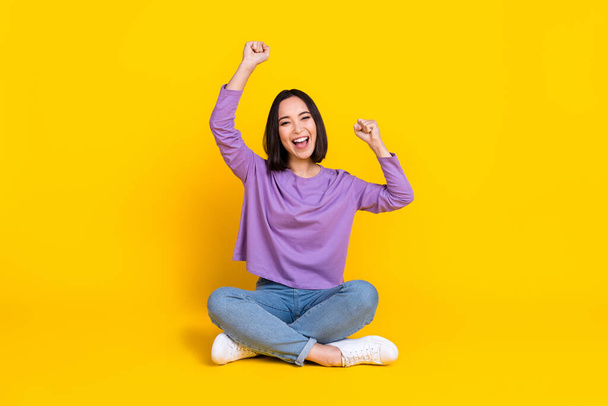 Photo of funny lucky lady dressed purple shirt sitting floor legs crossed rising fists isolated yellow color background. - Φωτογραφία, εικόνα