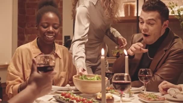 Lentitud media de alegres jóvenes y diversos amigos LGBTQ sentados en la mesa decorada con velas, bebiendo vino tinto y conversando. Anfitriona sirviendo ensaladas en cuencos - Imágenes, Vídeo