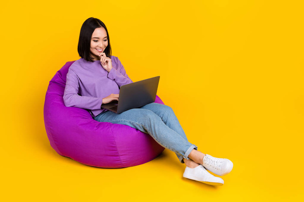 Full size photo of curious girl bob hairdo violet shirt pants sit on pouf look at laptop write email isolated on yellow color background. - Fotó, kép