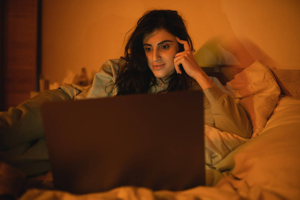 Bruna donna guardando il computer portatile offuscato sul letto di notte  - Foto, immagini