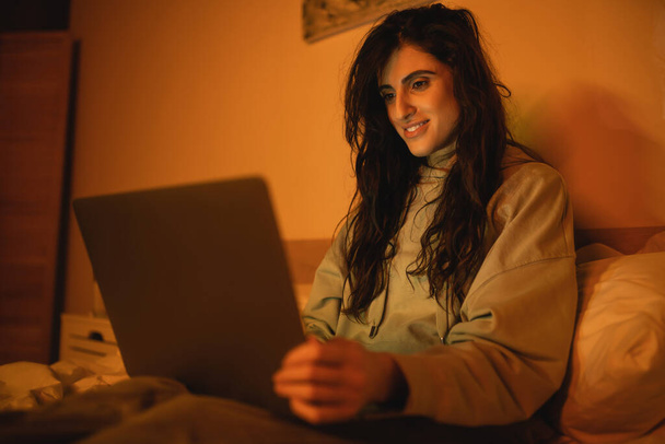 Mujer morena sonriente usando un ordenador portátil borroso en la cama por la noche  - Foto, Imagen