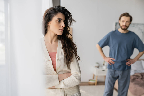 Angry woman crossing arms while standing near blurred boyfriend in bedroom  - Fotoğraf, Görsel