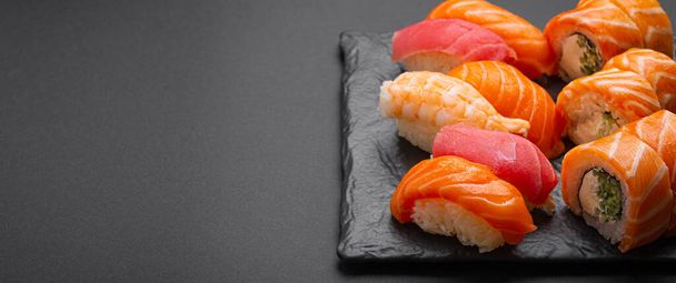 Set of traditional Japanese sushi and rolls angle view with soy sauce and wasabi on dark black rustic slate. Sushi with salmon, tuna and shrimp, space for text - 写真・画像