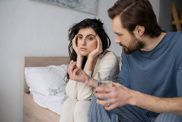 Caring man holding pills and water near frustrated girlfriend on bed at home  - Fotoğraf, Görsel