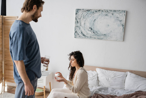Gondoskodó férfi kezében pohár víz közelében barátnője tabletták a hálószobában  - Fotó, kép