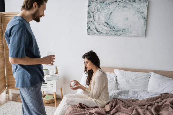 Vue latérale de la femme tenant des pilules près du petit ami attentionné avec un verre d'eau dans la chambre  - Photo, image