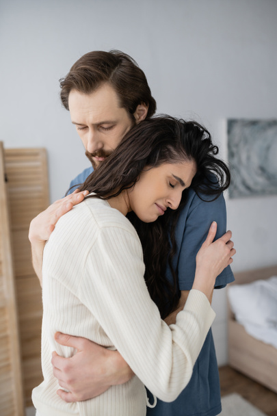 Bearded man hugging and calming displeased girlfriend in bedroom at home  - Photo, Image