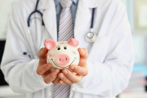 Beyaz önlüklü tıp doktoru mutlu domuz kumbarasını elinde tutuyor. Tıbbi bakım ekonomisi sağlık tasarrufu ve sigorta kavramı - Fotoğraf, Görsel