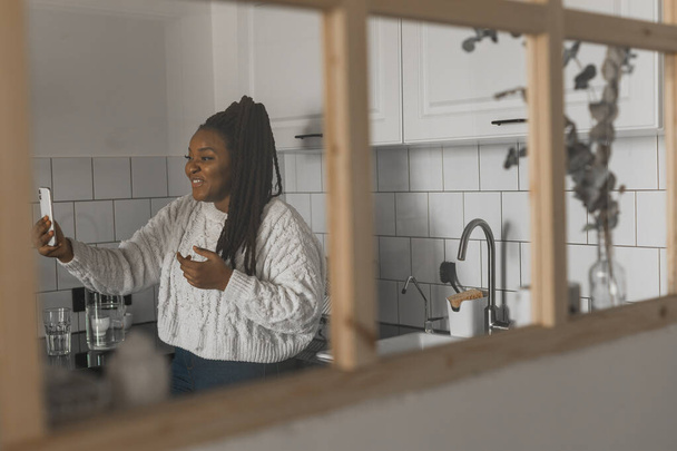 Happy modern African American young girl talking on video chat and sit home communication concept. Smiling hipster girl making self portrait on her telephone at home - Photo, image