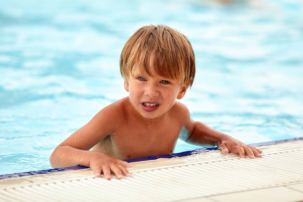 Vidám fiú a vízipark medencéjében, a gyerek boldogan pózol a parkban lévő kamerának a medence hátterében.. - Fotó, kép
