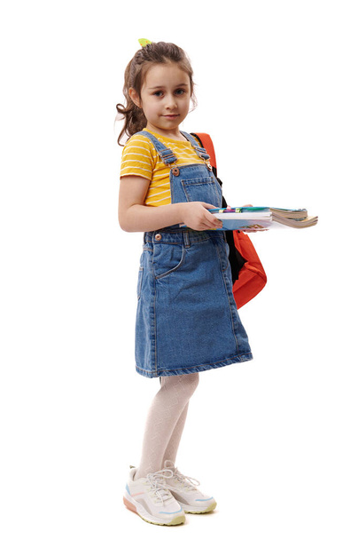 Pełny portret białej uśmiechniętej najlepszej uczennicy, uczennicy pierwszej klasy, 5-6-letniej dziewczynki trzymającej książki i książki do szkoły, odizolowanej na białym tle. Przestrzeń reklamy - Zdjęcie, obraz