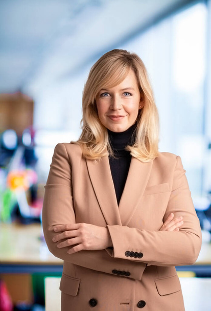 Close-up of mid aged businesswoman smiling and looking at camera. Blond haired female wearing blazer while standing at the office. Copy space.  - Foto, Bild