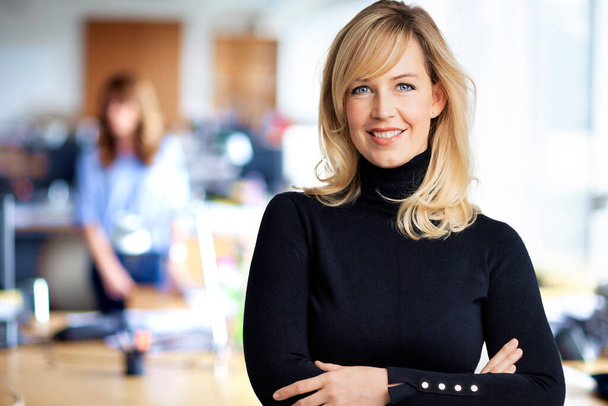 Close-up of mid aged businesswoman smiling and looking at camera. Blond haired female wearing black sweater while standing at the office. Copy space.  - Foto, afbeelding